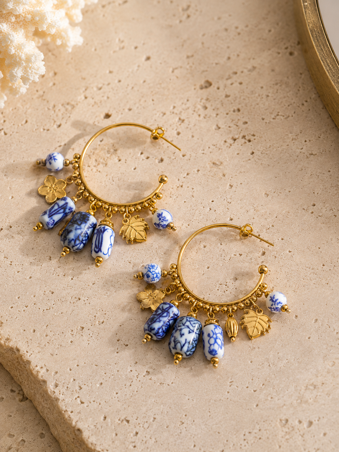 Women's open hoop drop earrings with blue and white ceramic beads, floral and leaf charms, PVD real gold plating and a layered statement design that highlights artisanal-inspired jewellery craftsmanship.
