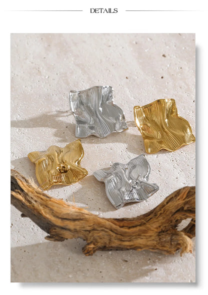 Four textured, abstract-shaped earrings in gold and silver tones arranged on a light beige stone surface with a piece of driftwood at the bottom. The word 'DETAILS' appears at the top.